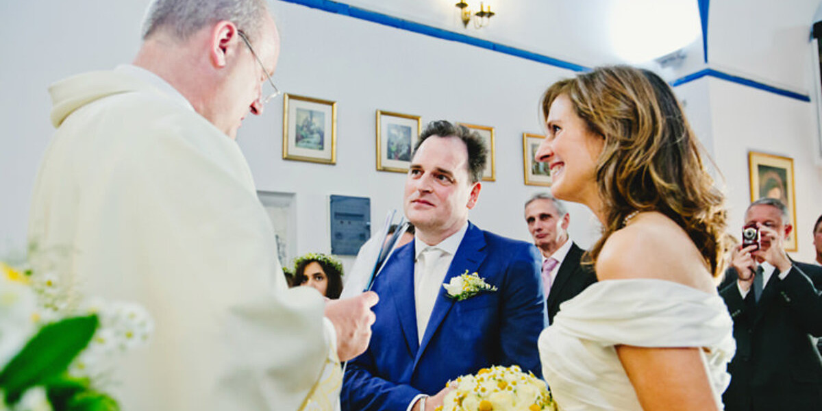 A couple stands before a priest during a Mykonos wedding ceremony in a Catholic church. The groom is in a blue suit, and the bride holds a bouquet of yellow and white flowers. Guests are in the background.