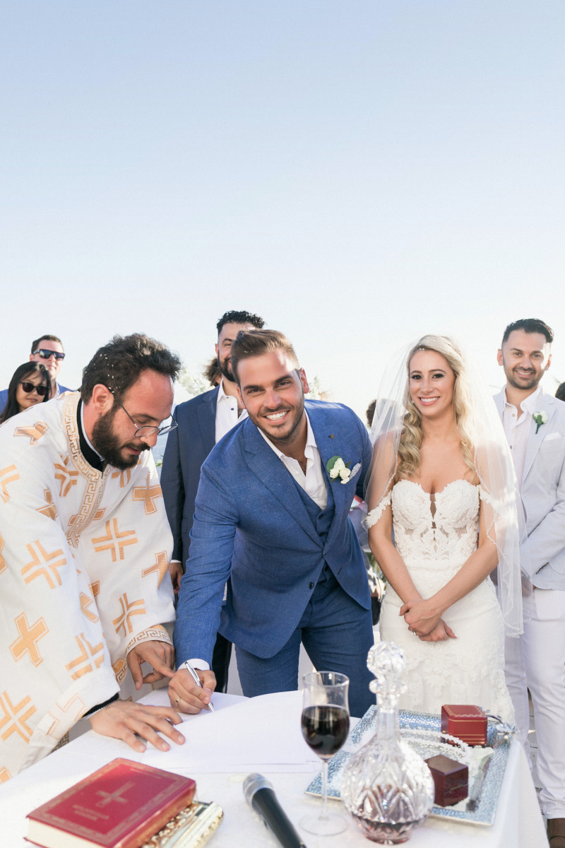 A newlywed couple in formal wedding attire signs a document at an outdoor ceremony, with guests and a Orthodox priest nearby.