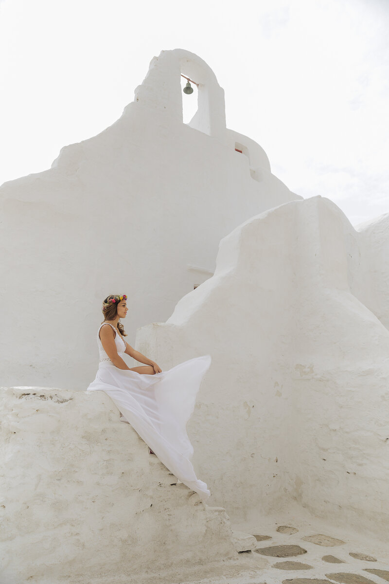 A woman in a flowing wedding white dress sits gracefully on a white stone wall, surrounded by rustic, whitewashed architecture under a cloudy sky at the traditional paraportiani in Mykonos.