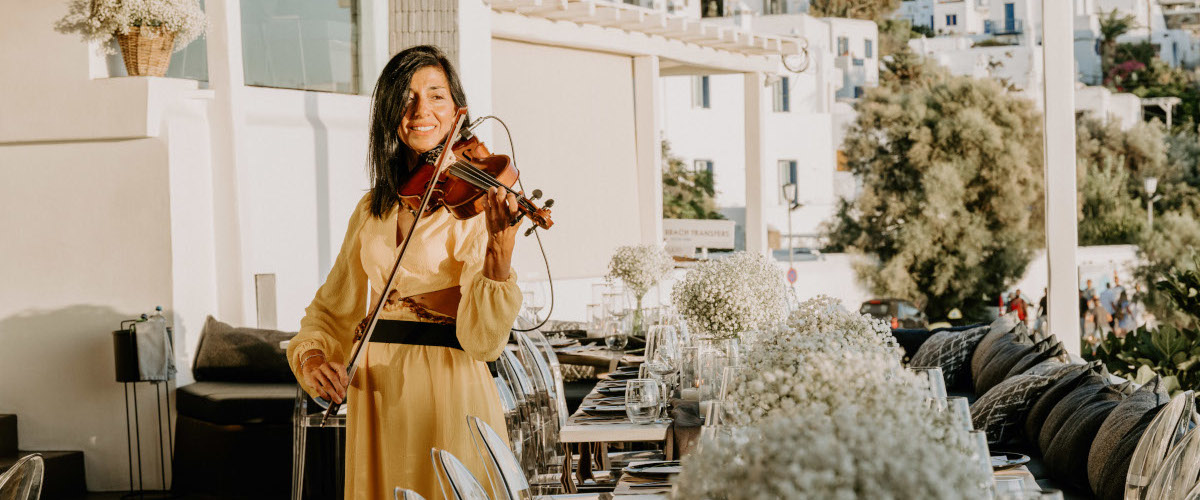 A woman in a bright yellow dress performs on the violin, along side to a decorated wedding dinner table in Mykonos island Greece